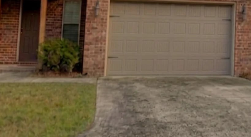 Brick house exterior with two-car garage door and driveway, landscaped lawn with shrub visible on left