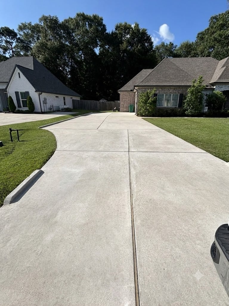 Before - Long concrete driveway leading to two residential houses with dark roofs, green lawns, and mature trees under blue sky